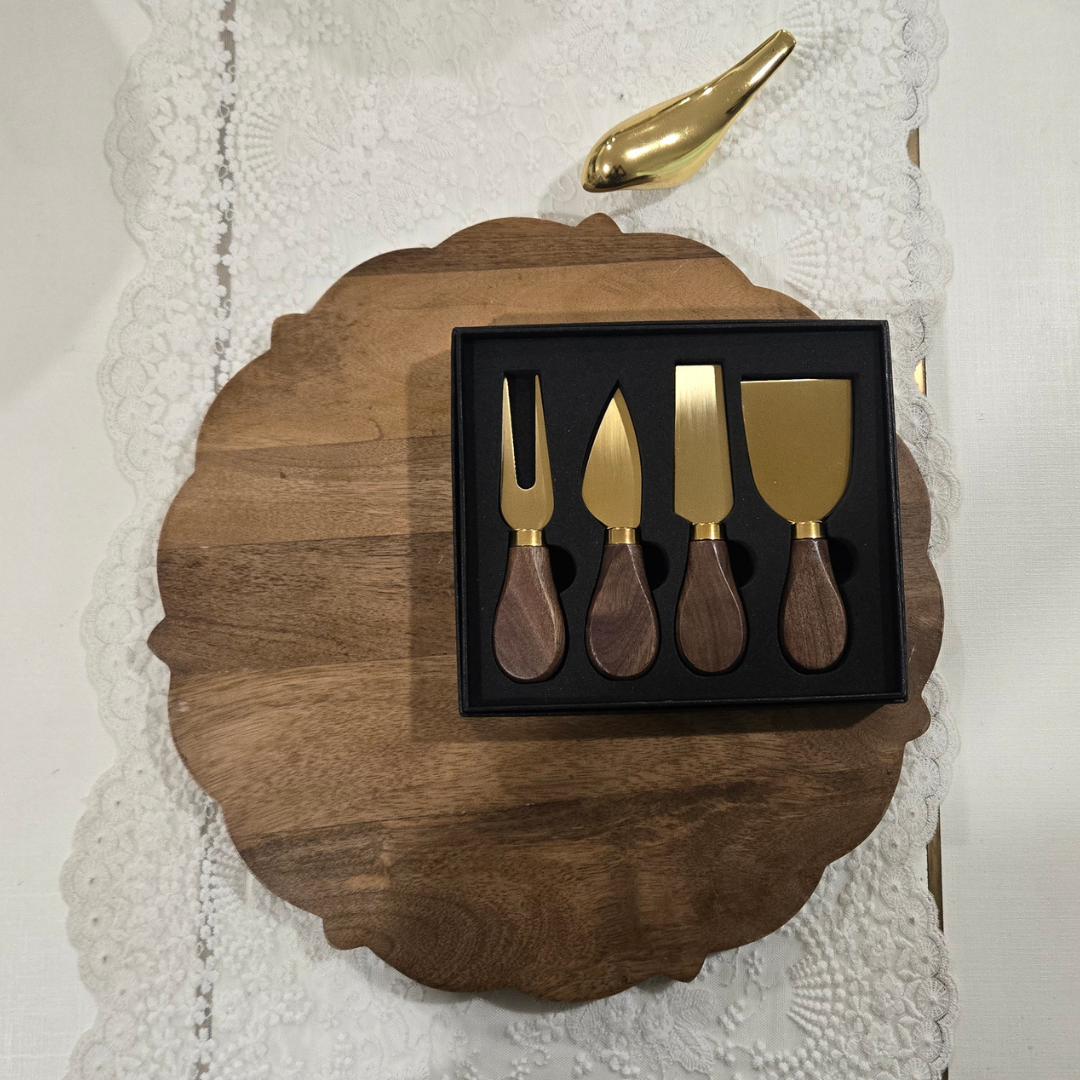 Four-piece cheese knife set with gold-tone metal heads and dark wood handles, presented in a black box on a scalloped wooden board over a white lace cloth.
