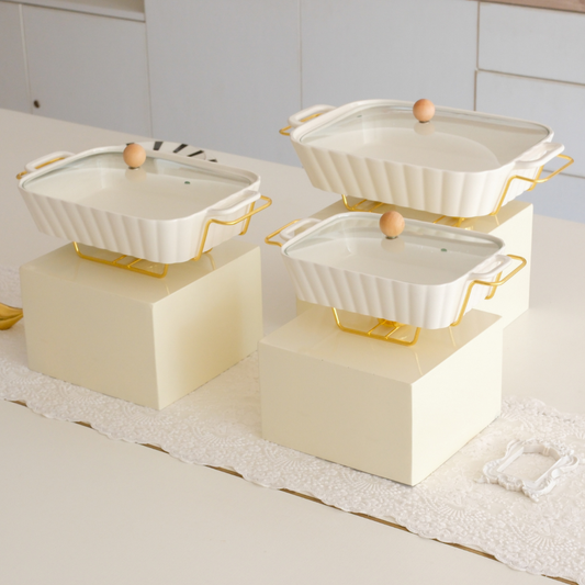 Four white ceramic casserole dishes with glass lids and gold-tone handles, displayed on cream-colored blocks on a kitchen counter.