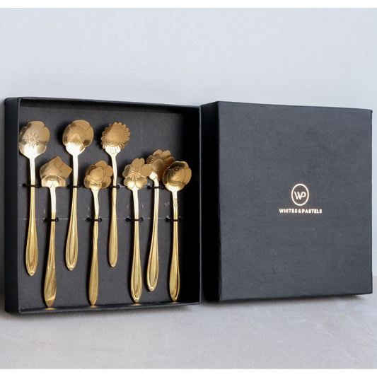 A set of eight gold-toned spoons with decorative floral and leaf-shaped spoils, presented in an open black gift box next to its lid, which features the White & Pastls logo.