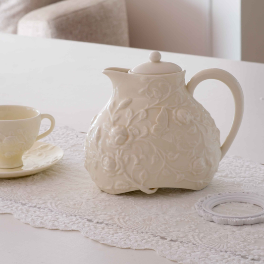 An off-white ceramic teapot with embossed floral patterns sits on a matching lace table runner on a white table, alongside a small matching teacup and saucer.