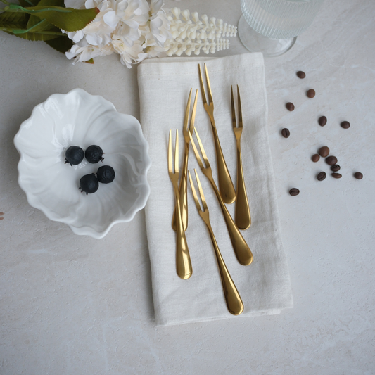 Six gold-toned flatware forks arranged on a folded cream linen napkin, styled on a light stone surface with a small white bowl of blueberries, scattered coffee beans, flowers, and a glass at the top.