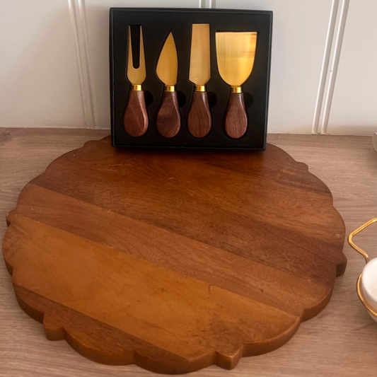 A wooden cheese knife set with gold-toned metal blades and dark brown wooden handles, displayed in a black gift box on a wooden surface with a large round wooden serving board in front.