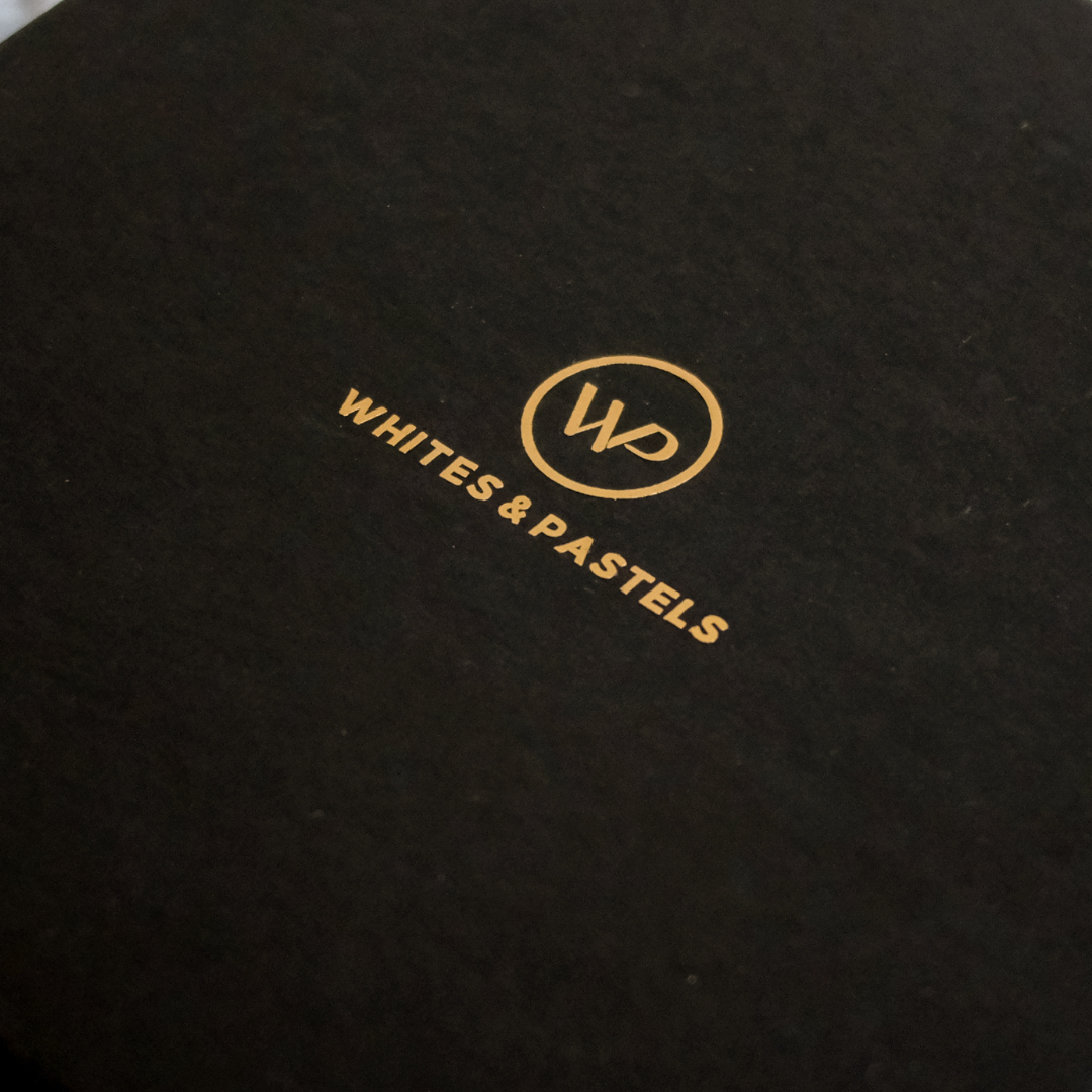 Close-up of a black textured surface with gold foil branding reading 'WHITES & PASTELS' and a circular 'WP' logo.