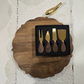 Four-piece cheese knife set with gold-tone metal heads and dark wood handles, presented in a black box on a scalloped wooden board over a white lace cloth.