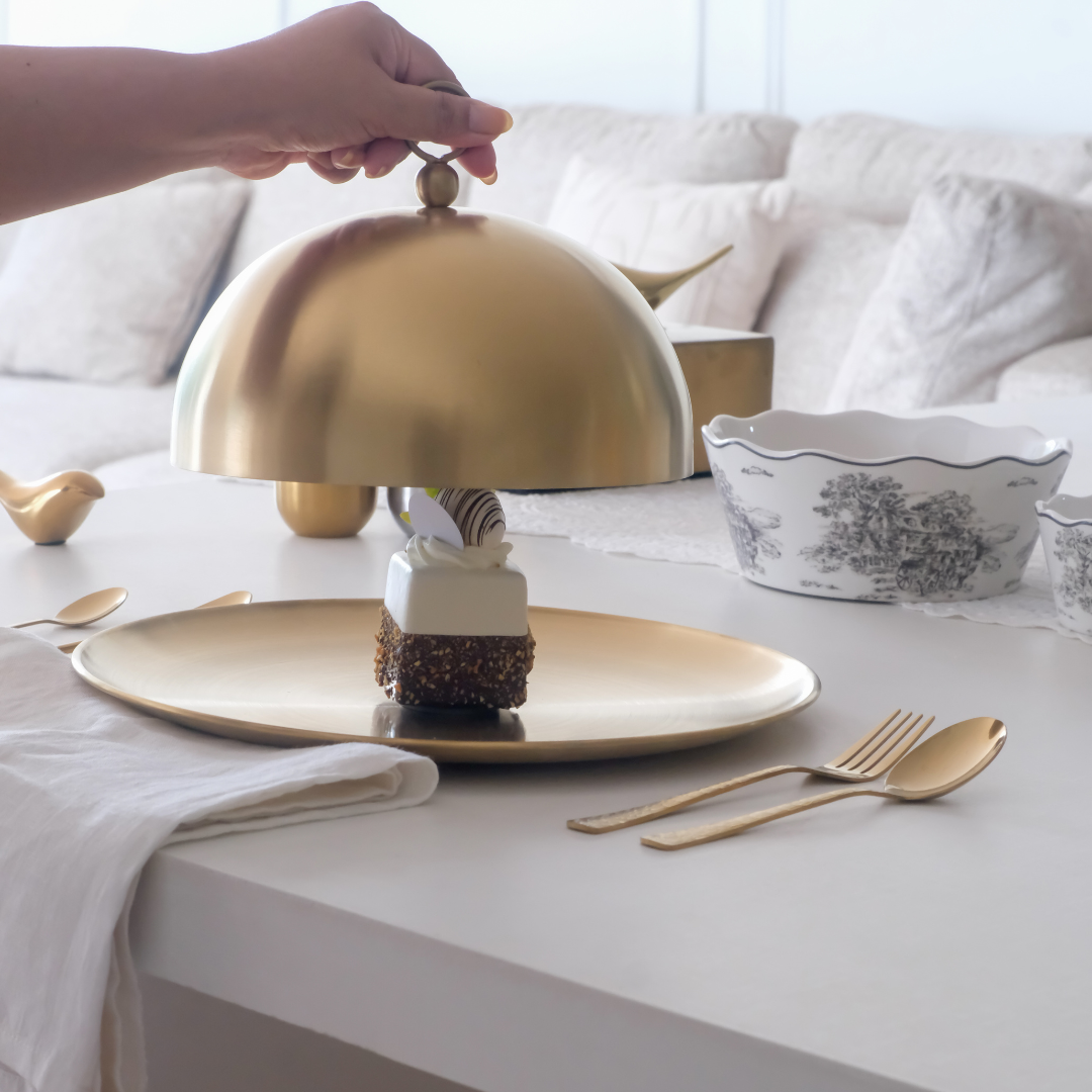 Gold cloche being lifted to reveal a dessert on a table with white and gold tableware.