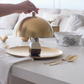 Gold cloche being lifted to reveal a dessert on a table with white and gold tableware.