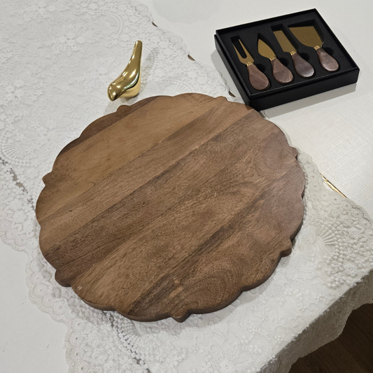 A round scalloped wooden serving board displayed on a white lace tablecloth, with a black gift box containing four gold-toned cheese knives and a small gold decorative object nearby.