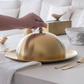 Gold cloche being held over a dish on a table with white background