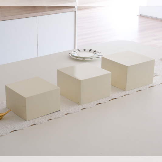 Three cream-colored cube side tables arranged on a light beige table runner in a bright, modern minimalist room.