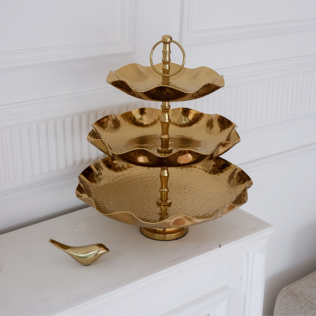 Three-tiered gold serving tray on a white surface with a white wall background