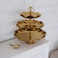 Three-tiered gold serving tray on a white surface with a white wall background