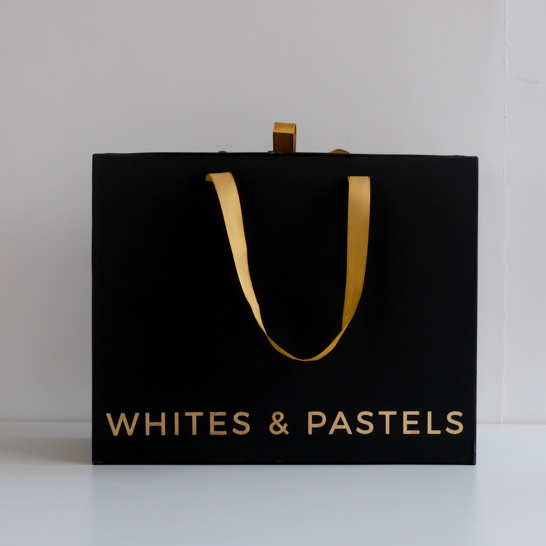 A black paper shopping bag with gold ribbon handles and gold foil lettering that reads 'WHITES & PASTELS' at the bottom, photographed against a plain light gray background.