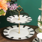 A two-tier cake stand with black and white stripes and a gold handle, displayed with a cake and cupcakes on it, set against a floral background.