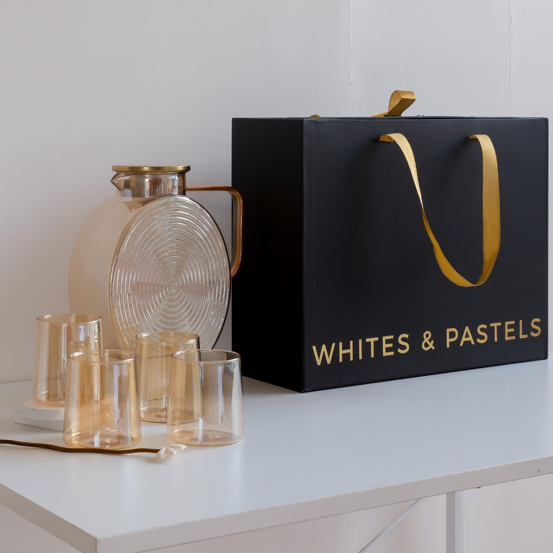 A set of amber glass drinking glasses and a matching glass carafe with gold-toned metal accents, displayed beside a large black gift bag printed with the brand name 'WHITES & PASTELS' and a gold ribbon handle, on a white tabletop against a white wall.