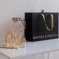 A set of amber glass drinking glasses and a matching glass carafe with gold-toned metal accents, displayed beside a large black gift bag printed with the brand name 'WHITES & PASTELS' and a gold ribbon handle, on a white tabletop against a white wall.