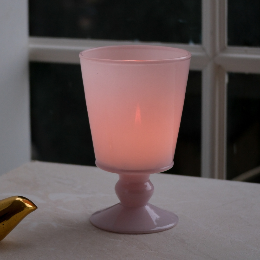 A pink frosted glass goblet candle holder with a pedestal base, illuminated from within, set on a light-colored stone surface with a window in the background.