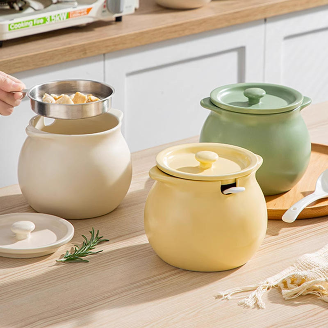 Three ceramic storage jars with lids in cream, pale yellow, and sage green, arranged on a light wood countertop in a kitchen setting. A hand is visible holding a metal bowl over the cream jar, and a white spoon rests on a wooden tray nearby.