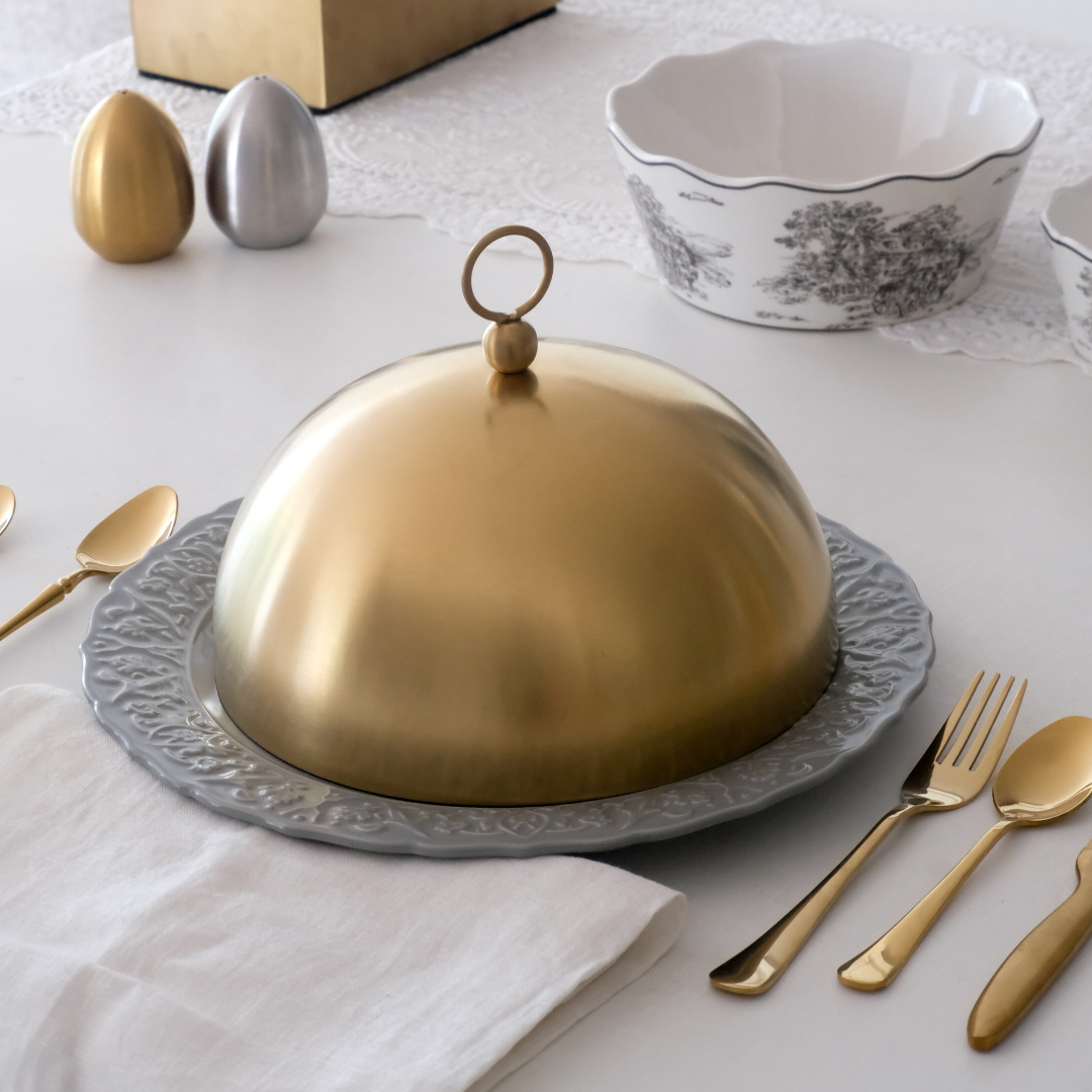 Gold cloche on a decorative platter with silverware and a bowl in the background