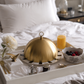 Tray with breakfast items including a glass of orange juice, bowl of berries, and gold-covered dish on a bed.