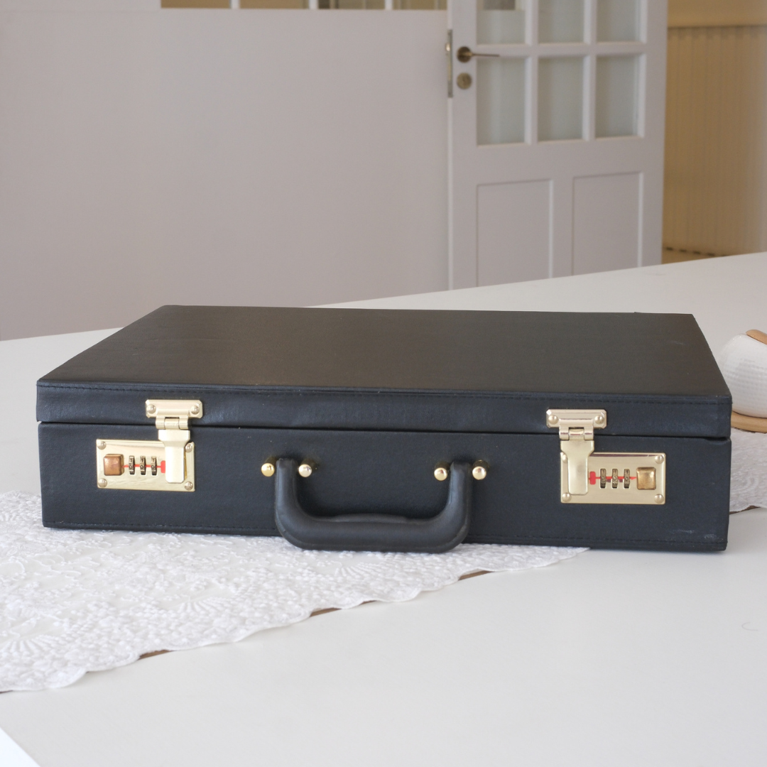 A black faux-leather briefcase-style storage box with gold-tone combination locks and a top handle, resting on a white table runner in a home setting.