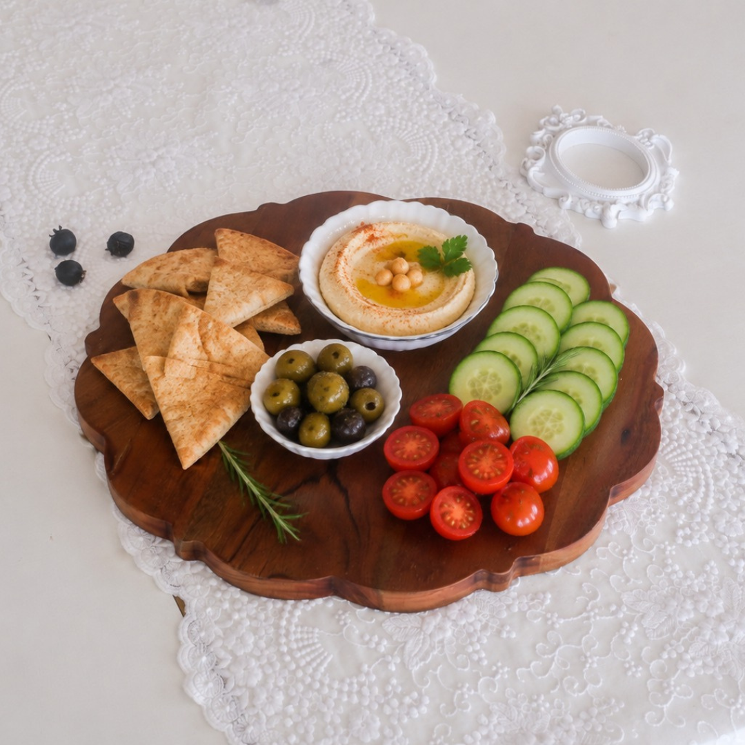 A scalloped wooden serving board styled with a Mediterranean charcuterie spread, including hummus, pita chips, olives, cucumber slices, cherry tomatoes, and blueberries, set on a white lace doily.