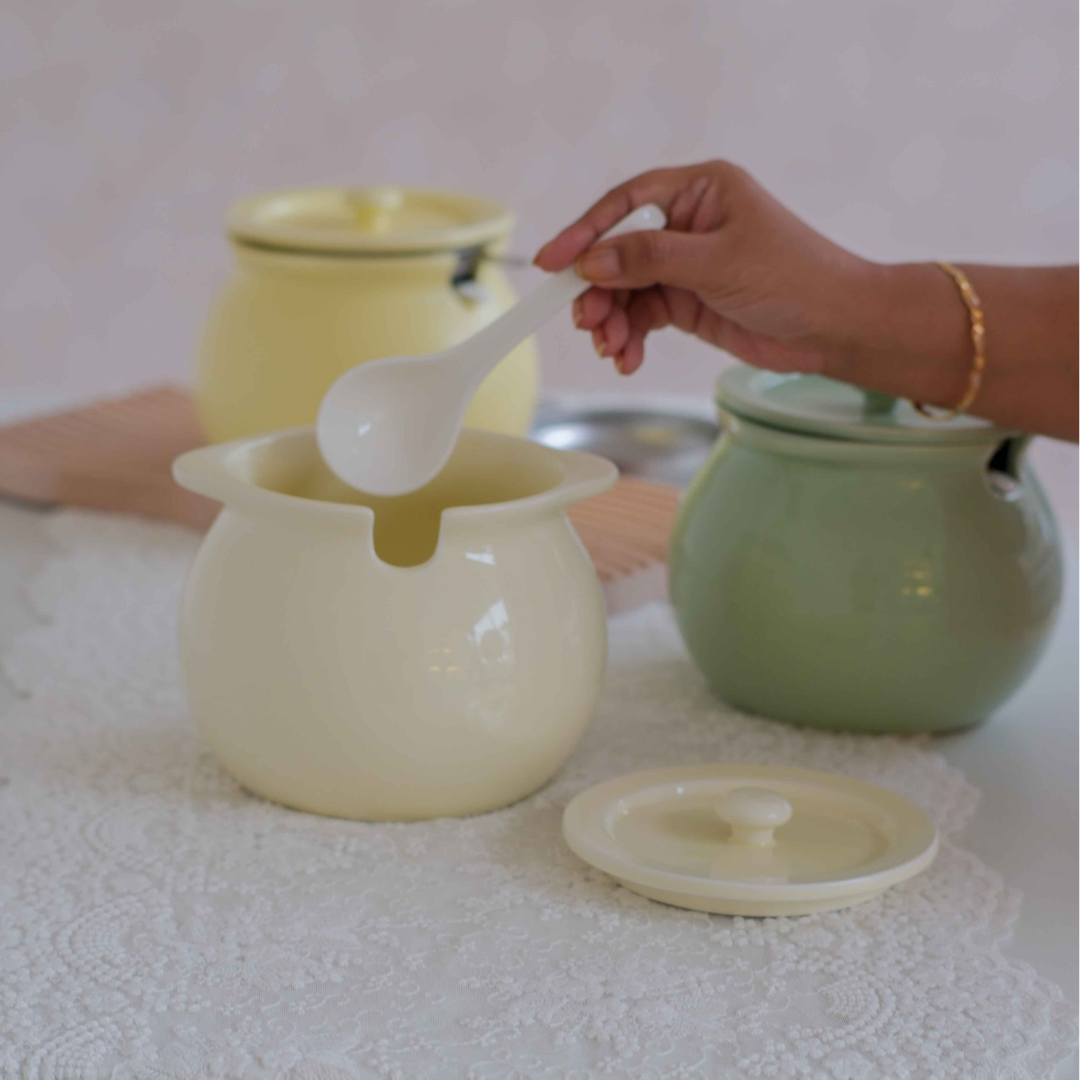 A ceramic sugar bowl in cream with a matching lid and white spoon, shown on a white lace tablecloth alongside other pastel-colored ceramic containers. A hand is holding the spoon.