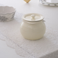 An off-white ceramic sugar ghee pot bowl with a matching lid and small side handle, placed on a white lace table runner. A patterned bowl is partially visible on the left, and other tableware appears softly blurred in the background.