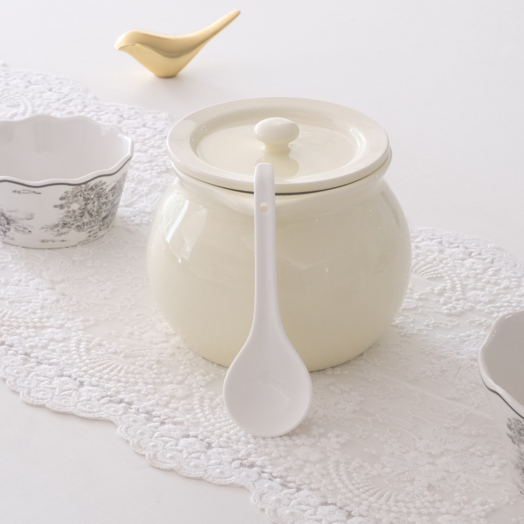 A cream-colored ceramic serving bowl with a matching lid and a white spoon resting in front, set on a white lace runner with other delicate tableware in the background.