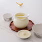 A ceramic tea set arranged on a white lace cloth, featuring a cream-colored teapot with a white spoon, a matching lid, a small cup, a bowl, and a wooden tray. A small gold bird figurine sits in the background.