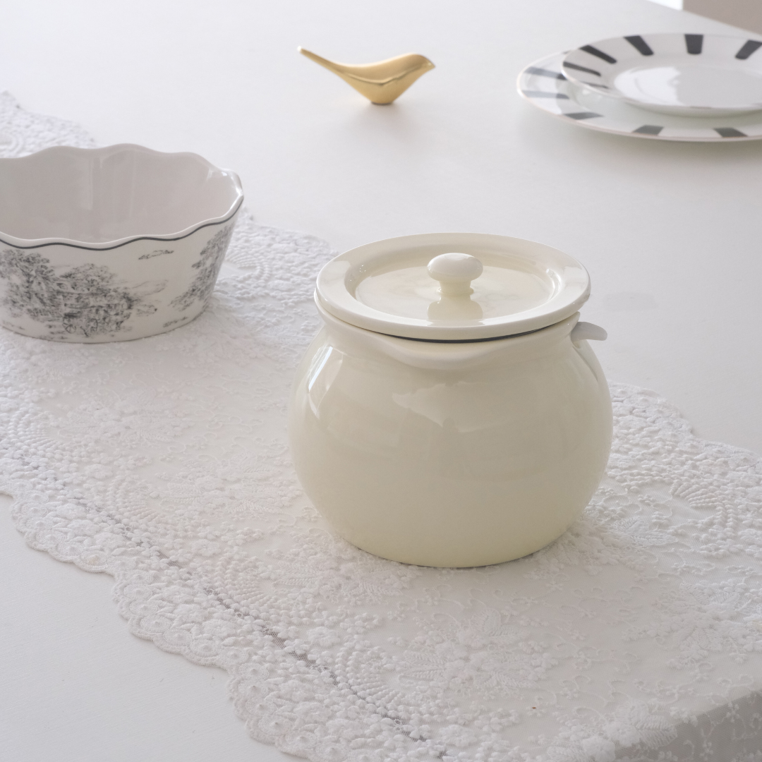 A white colored ceramic jar with a matching lid, placed on a white lace table runner on a white table. Other tableware, including a floral-patterned bowl and a small gold bird figurine, are visible in the background.