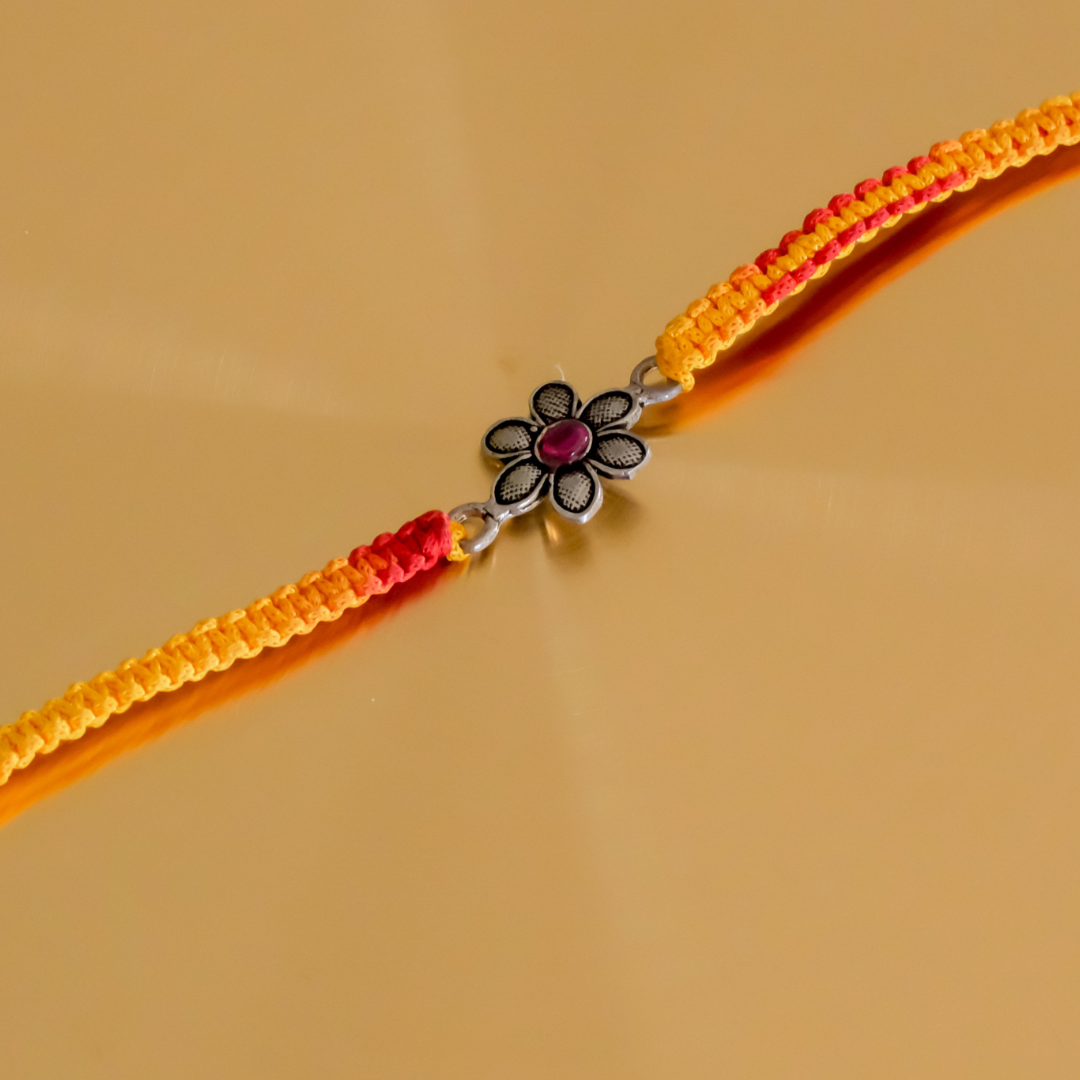Designer floral pure silver rakhi with intricate flower motif and pink gemstone on a handcrafted yellow-red thread for Raksha Bandhan.