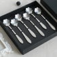 Six silver-toned metal spoons with flower-shaped heads and elongated handles, arranged inside a black presentation box on a light surface.