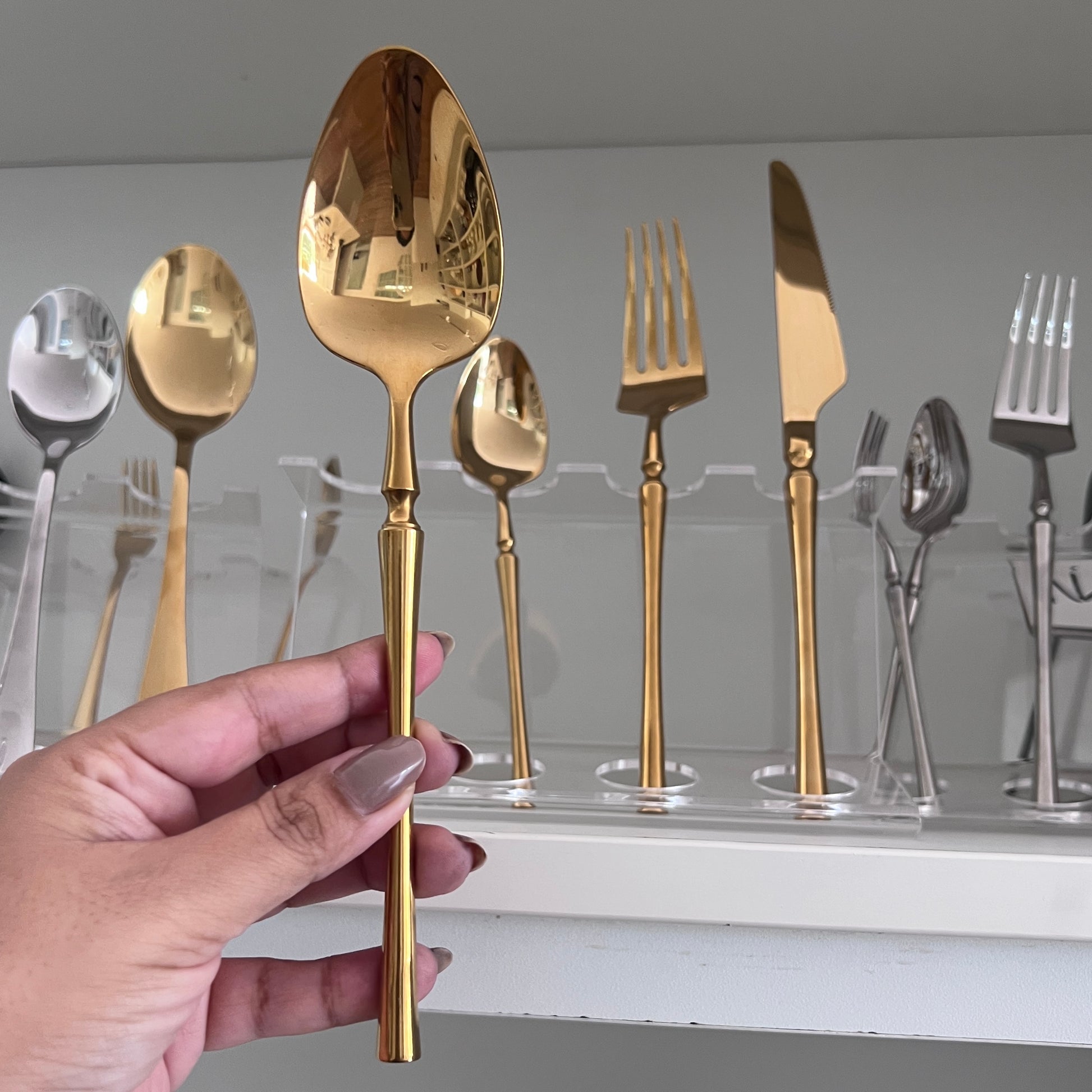 A hand holds a pvd finished gold spoon in the foreground, with a set of gold and silver flatware displayed on clear stands in the background.