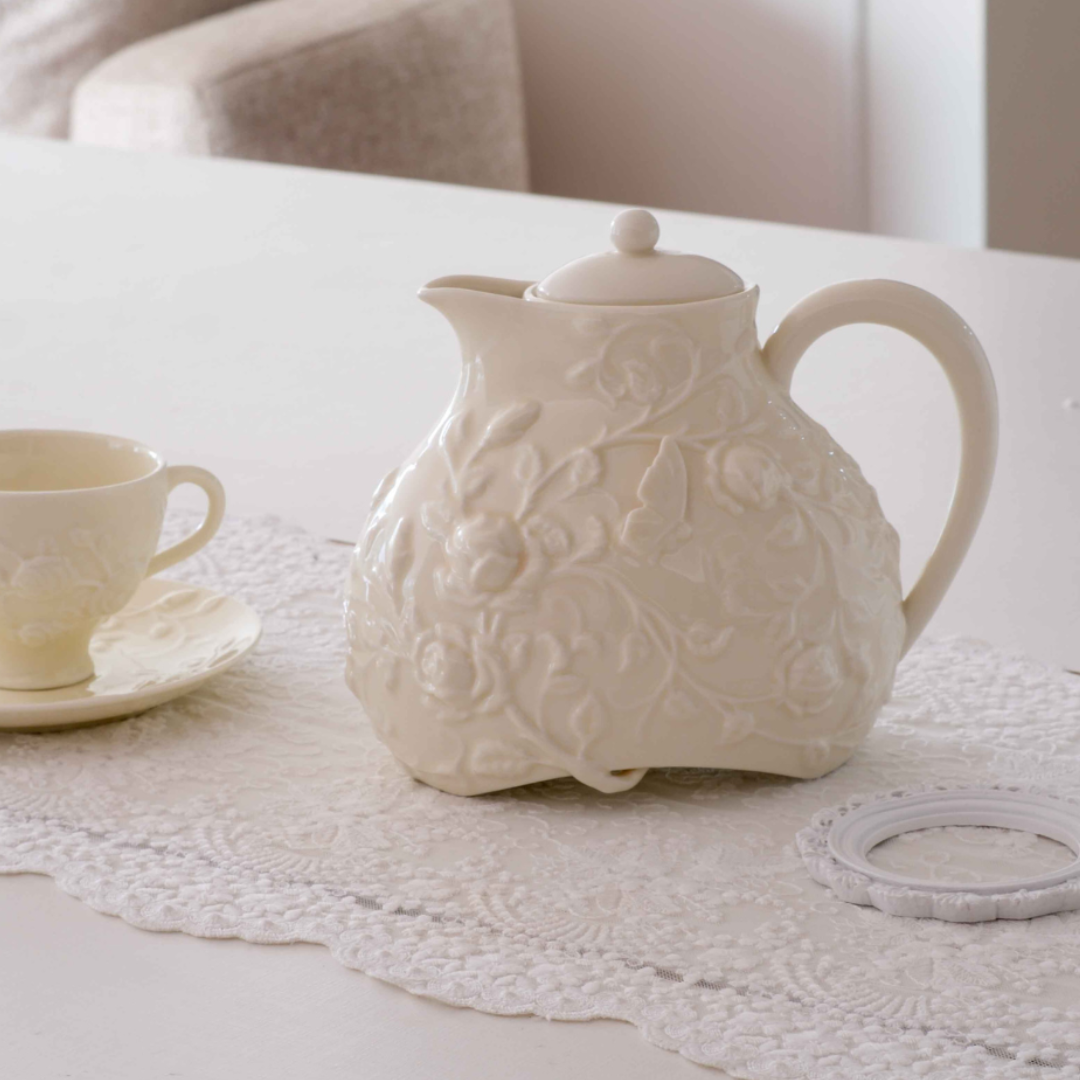 An off-white ceramic teapot with embossed floral patterns sits on a matching lace table runner on a white table, alongside a small matching teacup and saucer.