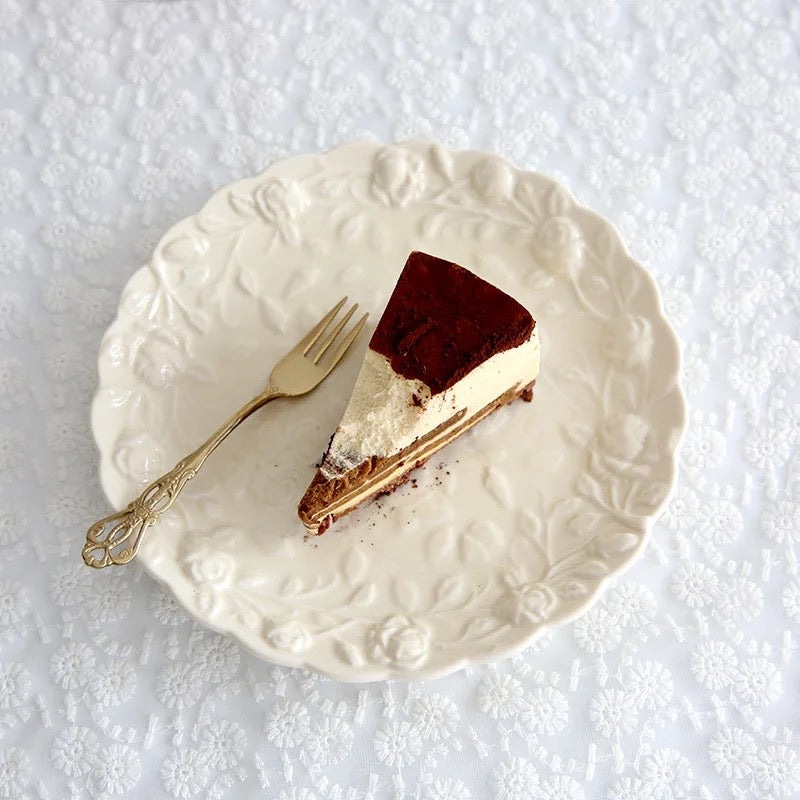 A slice of cake on an embossed white porcelain snack plate with a fork on the side, the plate has a floral like pattern around the edge. Color of plate is ivory.