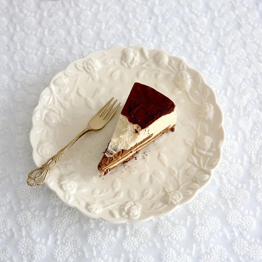 A slice of cake on an embossed white porcelain snack plate with a fork on the side, the plate has a floral like pattern around the edge. Color of plate is ivory.