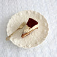 A slice of cake on an embossed white porcelain snack plate with a fork on the side, the plate has a floral like pattern around the edge. Color of plate is ivory.