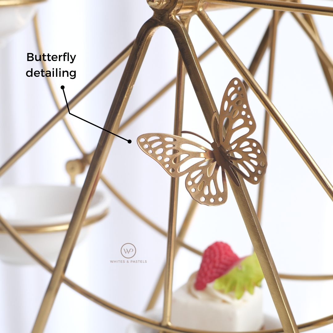 Close-up of a gold metal food display stand with a butterfly-shaped ornament attached to one of its arms. The image includes a text callout reading 'Butterfly detailing and a small logo near the bottom that say Whites & Pastels.