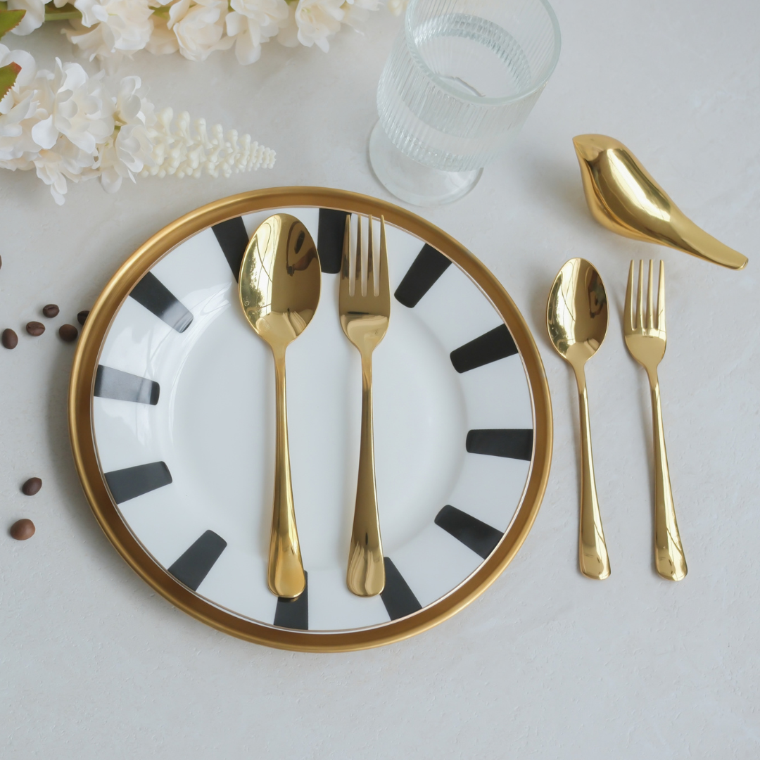 Gold PVD dinnerware set including a spoon, fork, and knife, arranged on a white plate with black stripes and a gold rim, alongside a gold bird-shaped object on a light gray surface.