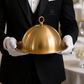 Waiter in formal attire holding a gold cloche dish with a blurred background