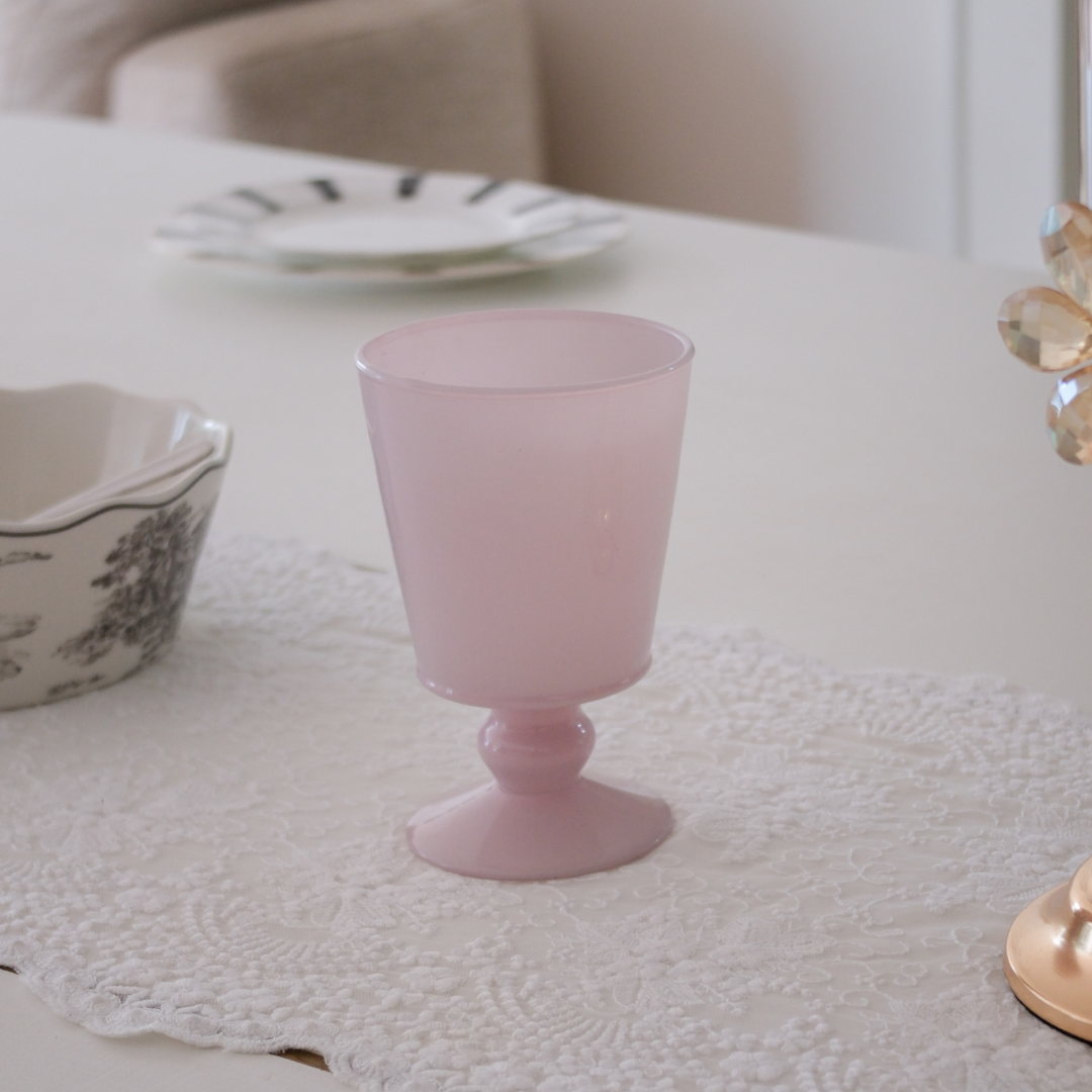 A light pink frosted glass goblet with a flared base, placed on a white lace table runner on a dining table.