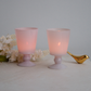 Two pink frosted glass candle holders with lit candles inside, styled on a light surface with white flowers and a small gold decorative bird on a neutral background.