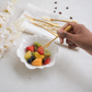 A hand holds a gold-toned fork over a small white scalloped bowl filled with sliced fruit, with additional gold forks and a white napkin in the background on a light surface.
