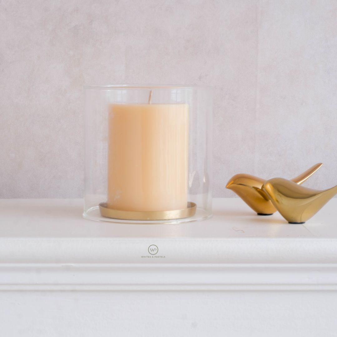 Candle in a glass holder with gold base on a white surface, next to gold decorative shoes.
