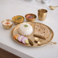 A gold-toned round serving platter styled with a mound of white rice, folded flatbread, sliced red onions, lime, and carrots, alongside three small gold bowls of curry and a gold cup on a white tabletop.
