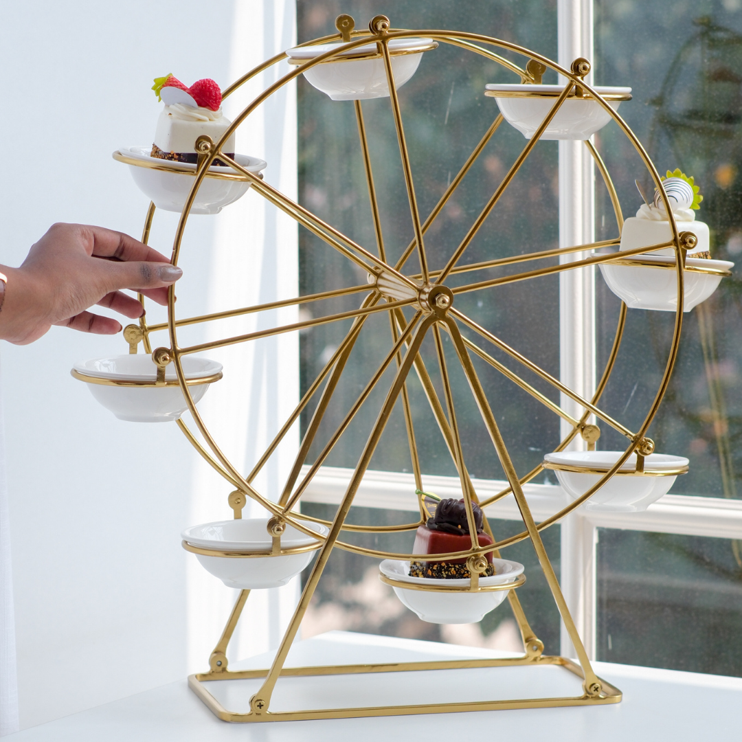 A gold-finished Ferris wheel dessert stand with white ceramic bowls, shown on a windowsill with a hand adjusting one of the bowls. Pastries are placed in several of the bowls.