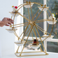 A gold-finished Ferris wheel dessert stand with white ceramic bowls, shown on a windowsill with a hand adjusting one of the bowls. Pastries are placed in several of the bowls.