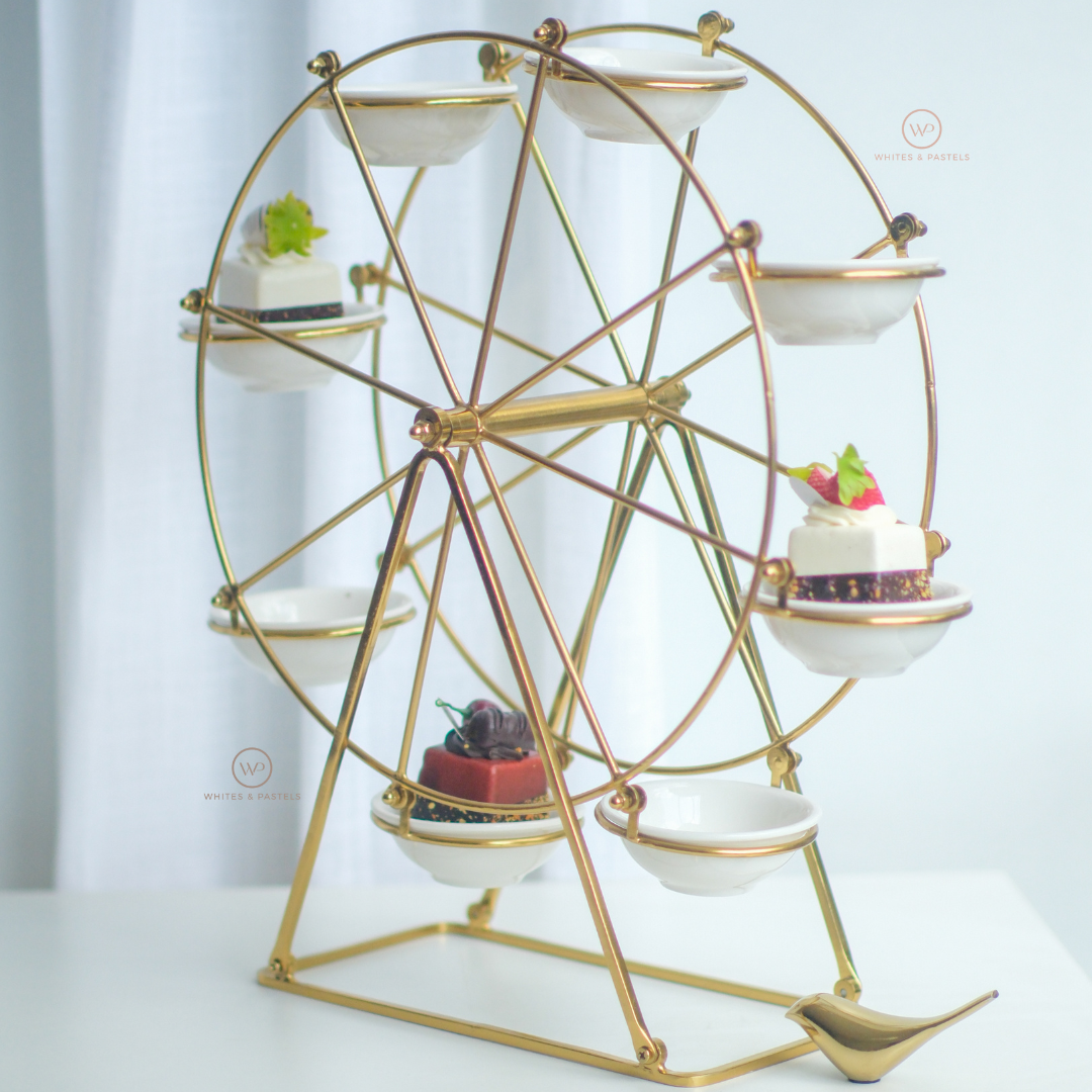 A gold metal Ferris wheel serving stand with eight white ceramic cups, styled with desserts, on a white table against a soft white curtain background.
