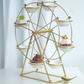 A gold metal Ferris wheel serving stand with eight white ceramic cups, styled with desserts, on a white table against a soft white curtain background.