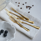 Six gold PVD stainless steel fruit forks arranged on a white linen napkin, with scattered coffee beans and a small white dish of blueberries in the background.